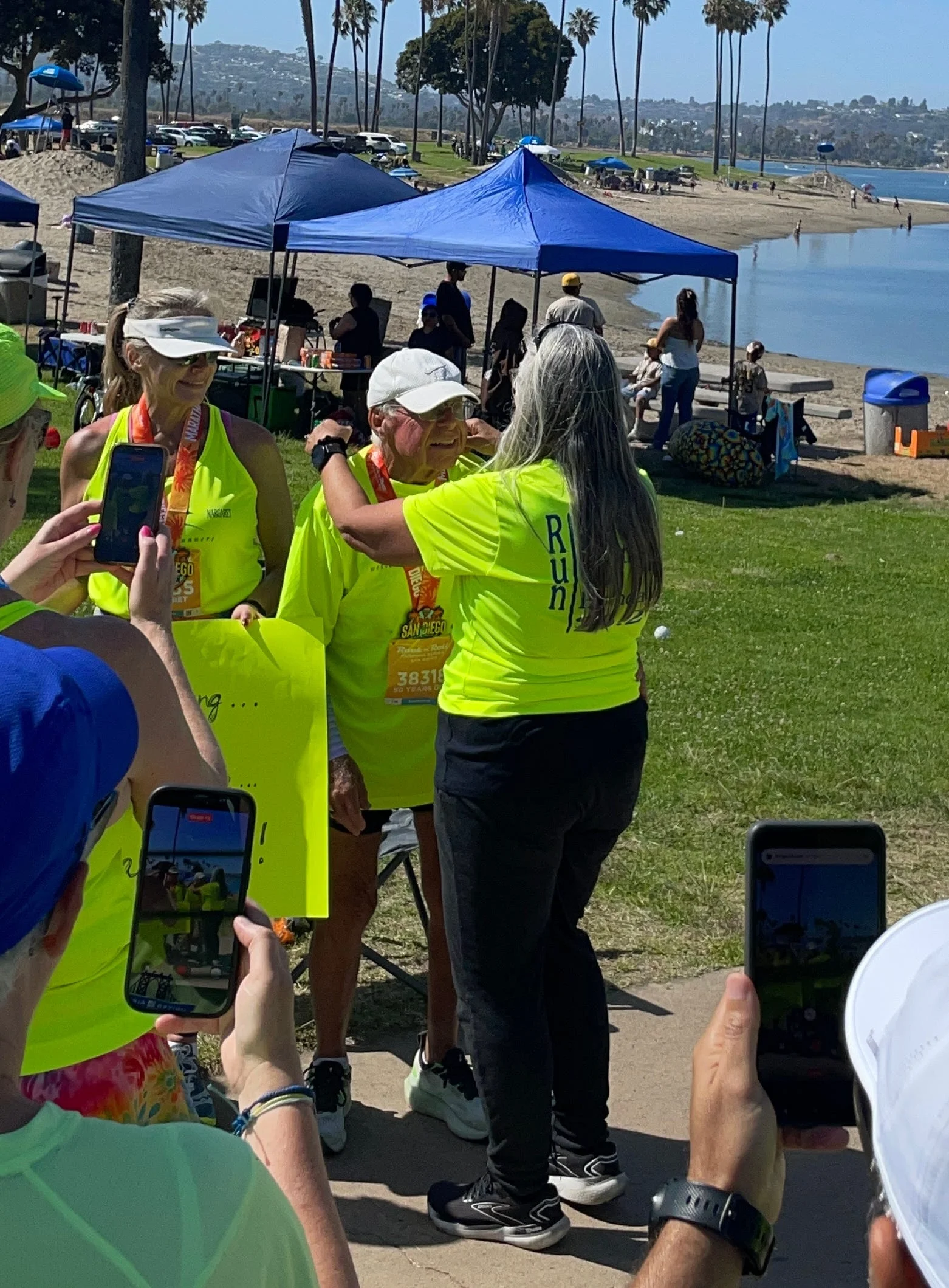 Runners in bright shirts celebrate at a beachside event, capturing memories with smartphones in sunny weather.