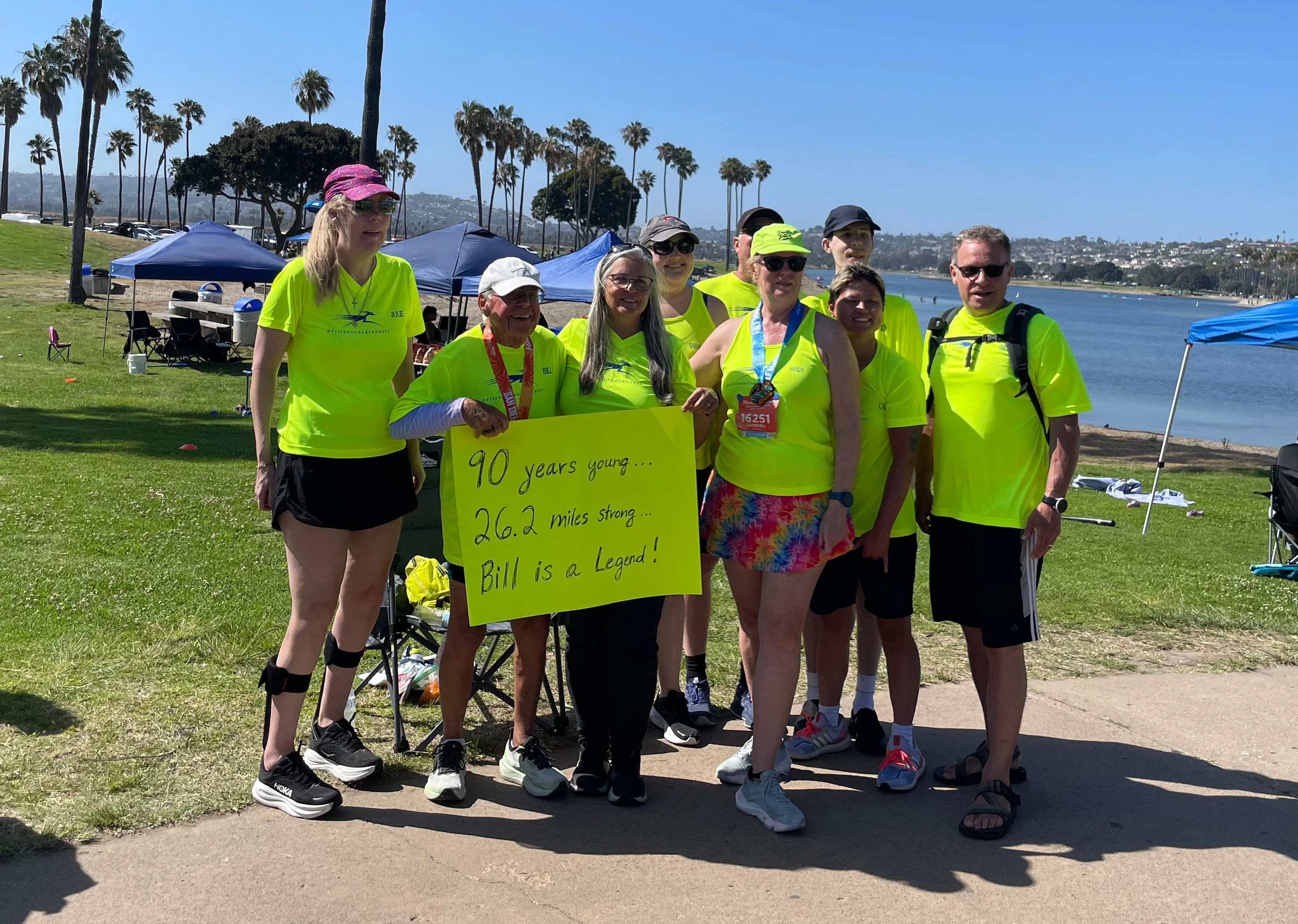 Group of runners celebrating a 90-year-old participant, holding a sign that recognizes his achievement.