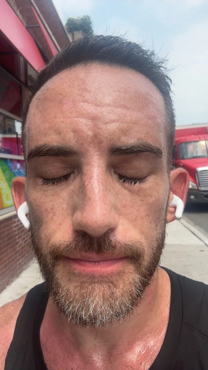 Man with closed eyes and earbuds, showing a focused expression, with a slightly sweaty face and a brick background.