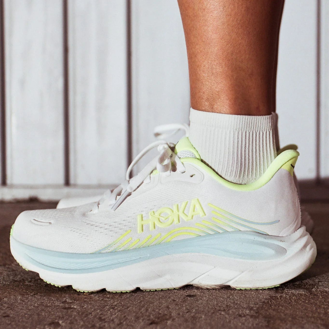 HOKA running shoes with a white and neon green design, worn on a person's feet against a textured surface.