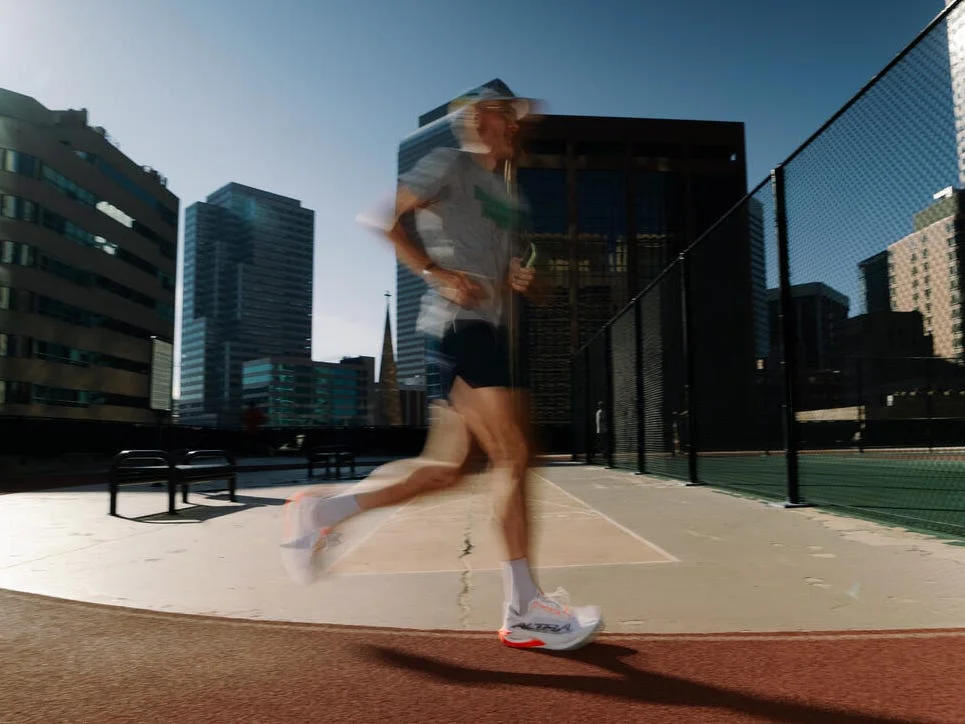 Blurred motion of a runner on a track with city buildings in the background and bright sunlight.