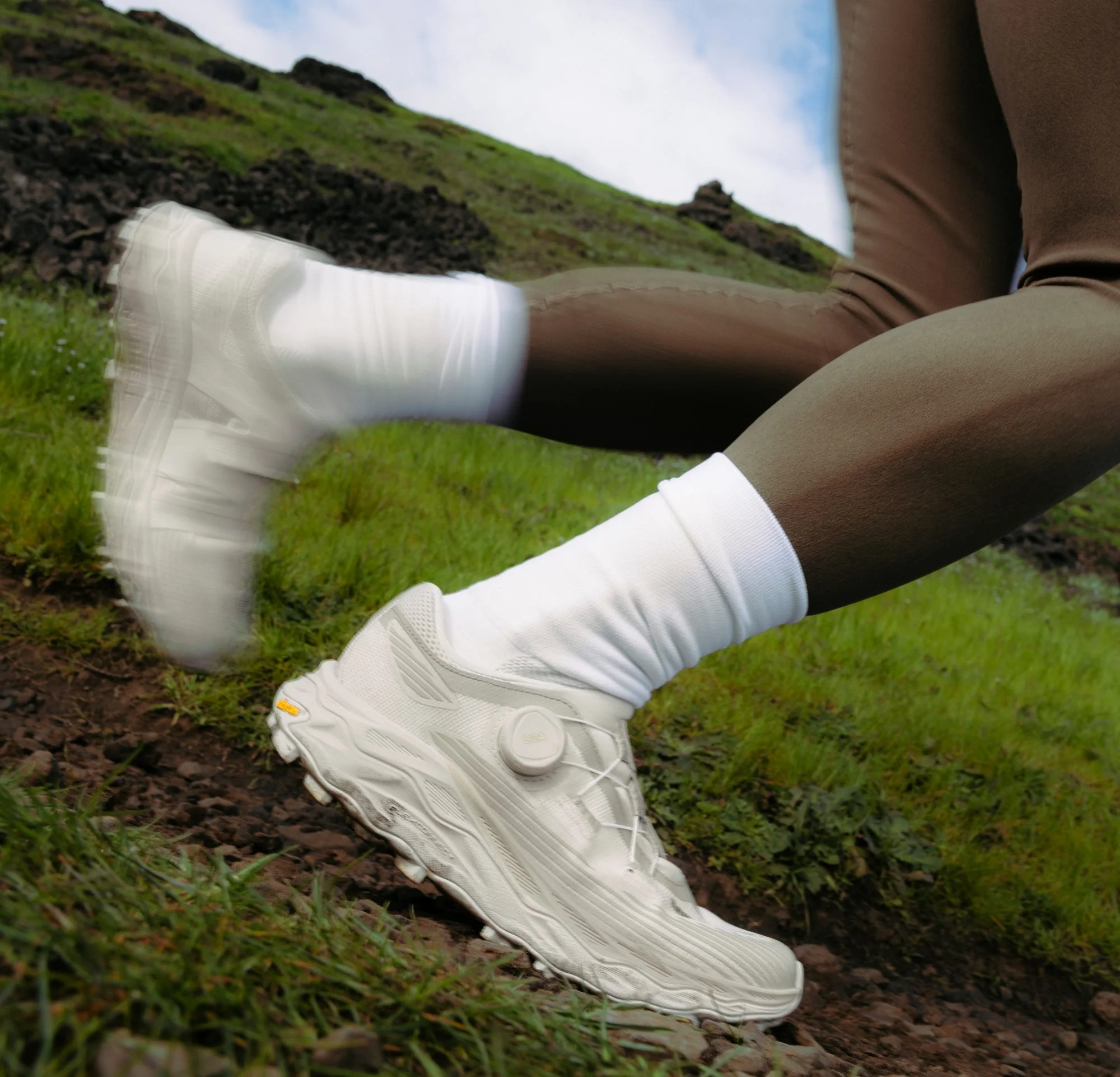 Person running on a grassy trail, wearing white sneakers and socks with brown leggings against a blue sky.