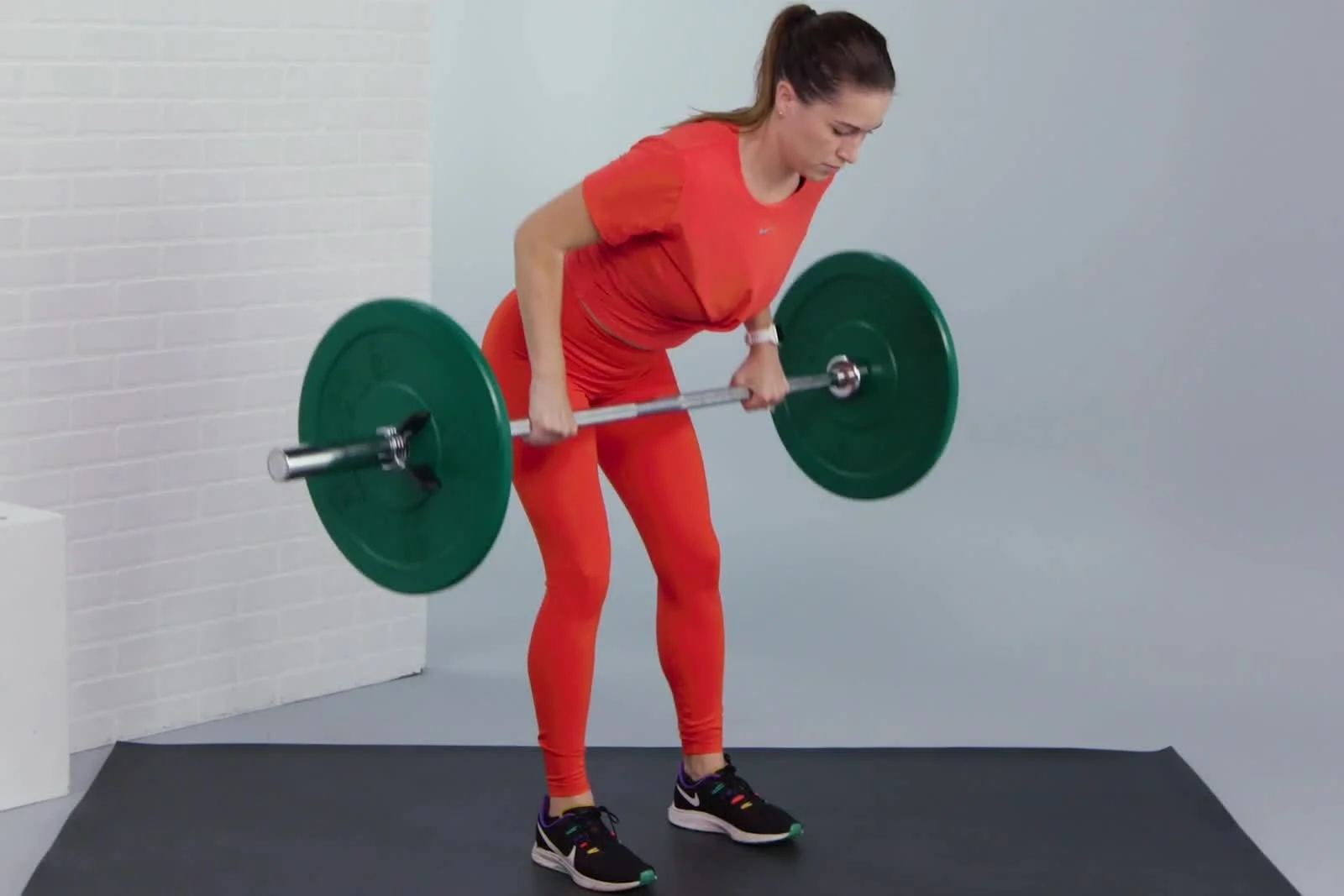 A person in red athletic wear performs a bent-over row exercise with a barbell on a gym mat.