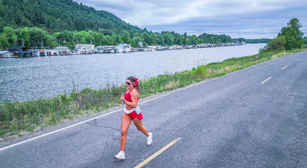 A person jogging along a road by a river with houses and lush greenery in the background on a cloudy day.