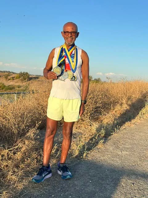 An older athlete proudly displaying multiple medals while standing on a gravel path with dry grass in the background.