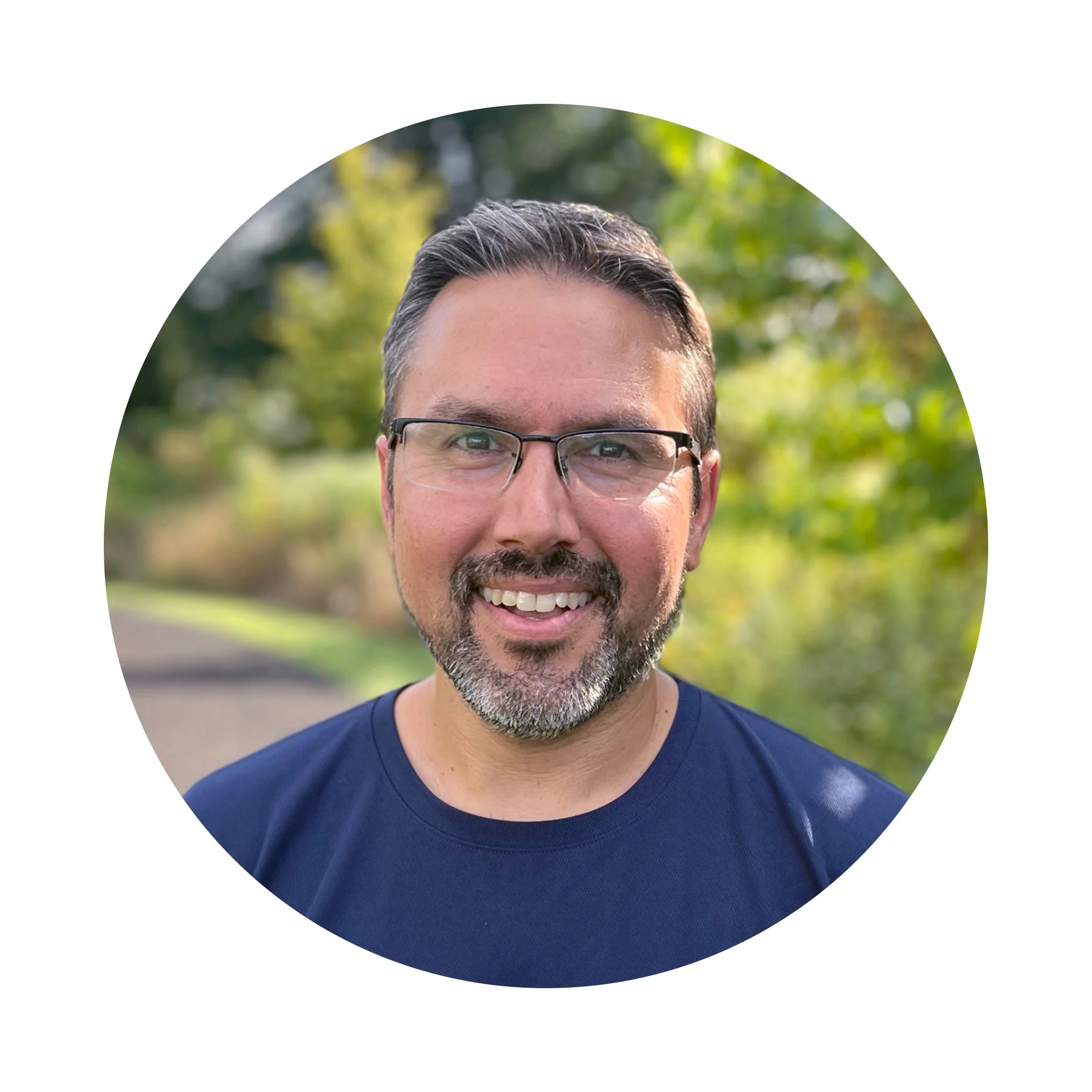 Smiling man with glasses and a beard, wearing a navy shirt, standing outdoors with greenery in the background.