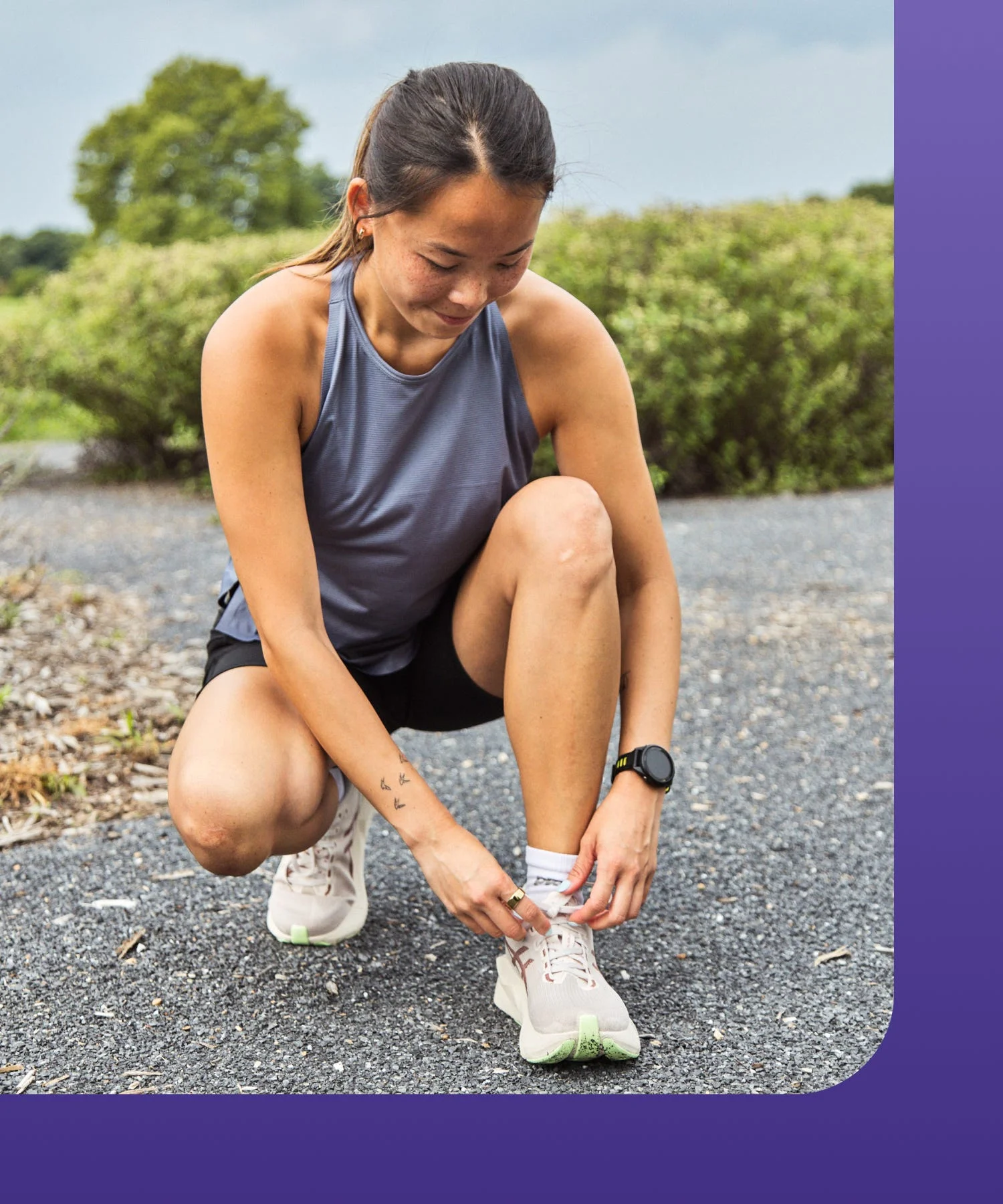 A woman crouches tying her running shoes on a gravel path with greenery in the background.