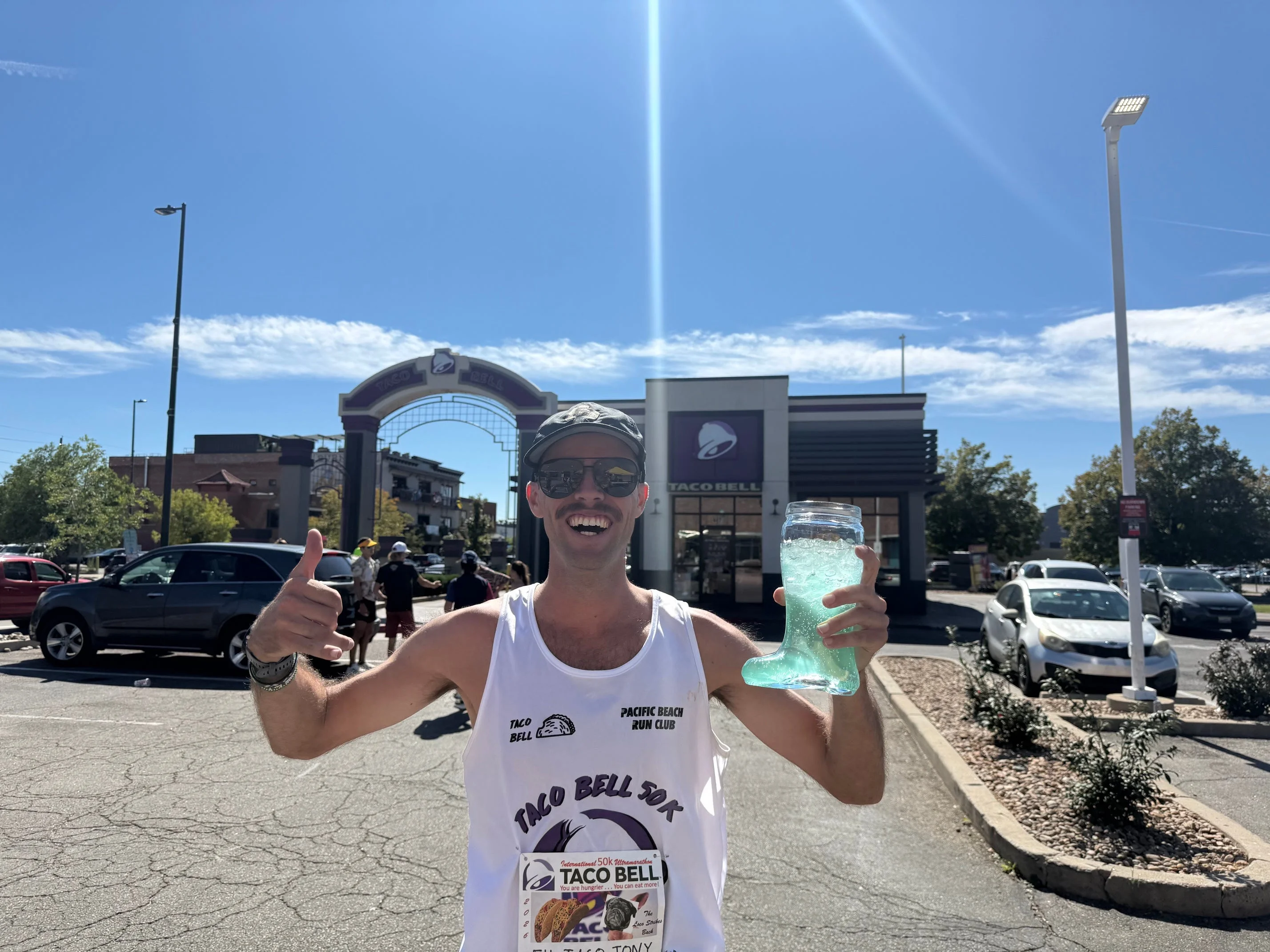 Man giving a thumbs up while holding a large drink in front of a Taco Bell restaurant on a sunny day.
