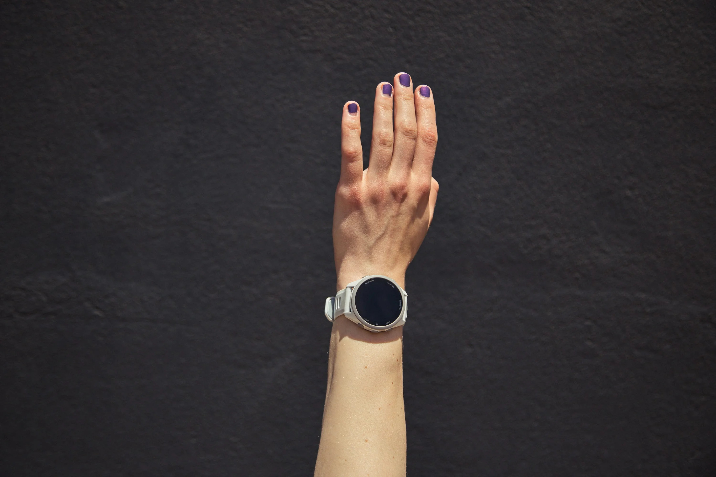 Hand with purple nails wearing a smartwatch against a dark background.