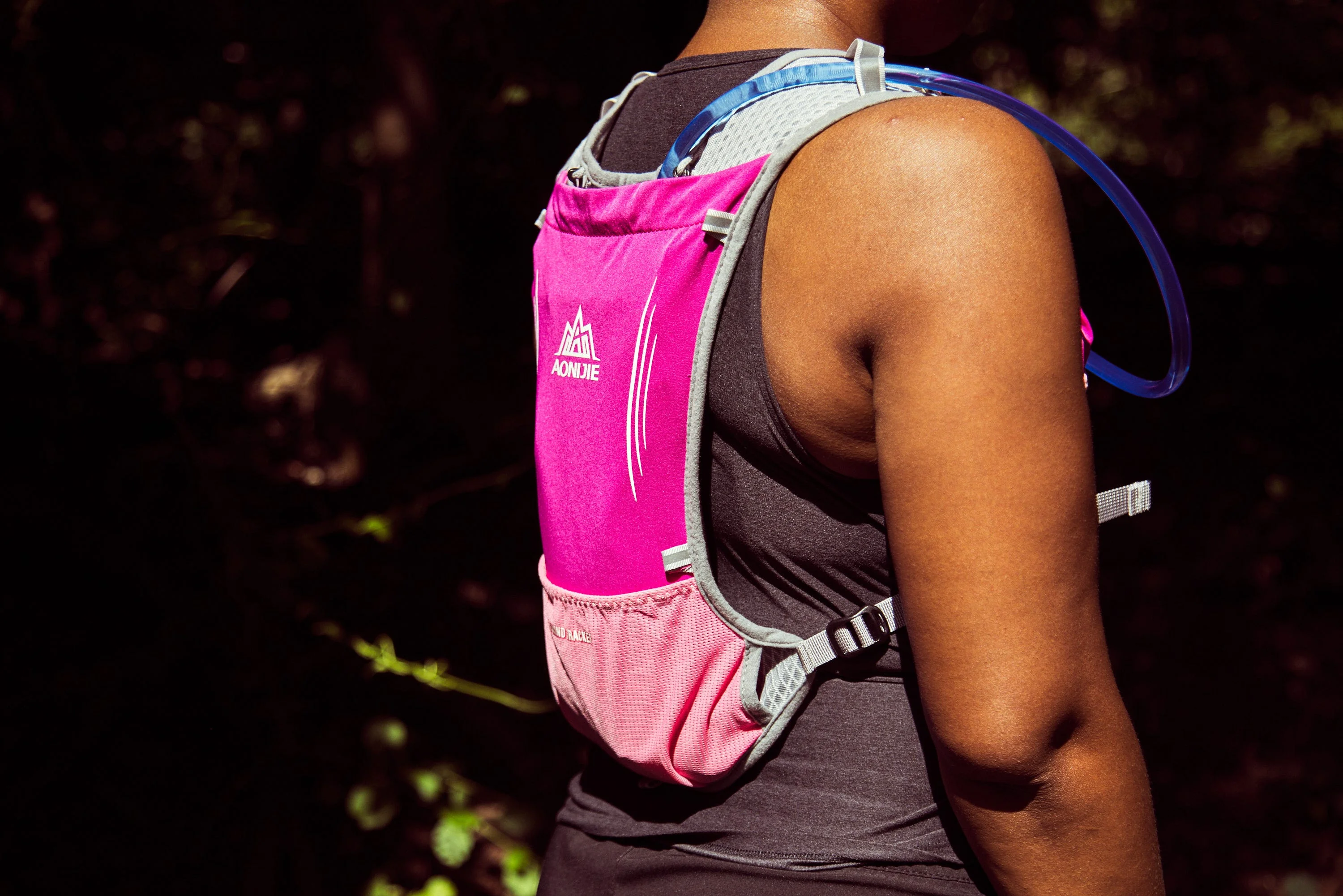 A close-up of a person wearing a pink hydration pack with a drinking tube in a forested setting. A close-up of a person wearing a pink hydration pack with a drinking tube in a forested setting.