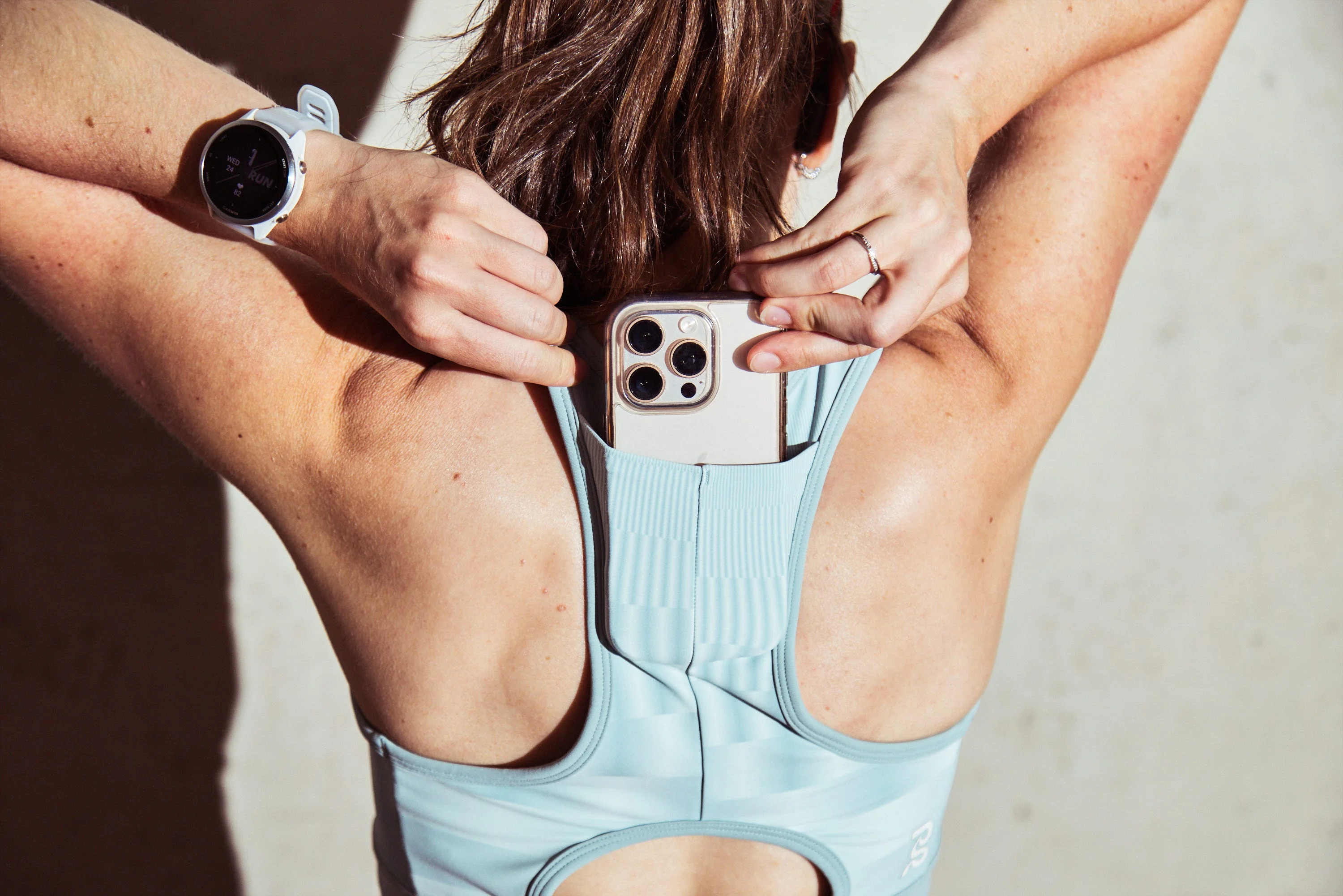 A person securing a smartphone in the back pocket of a light blue athletic top while wearing a fitness watch. A person securing a smartphone in the back pocket of a light blue athletic top while wearing a fitness watch.