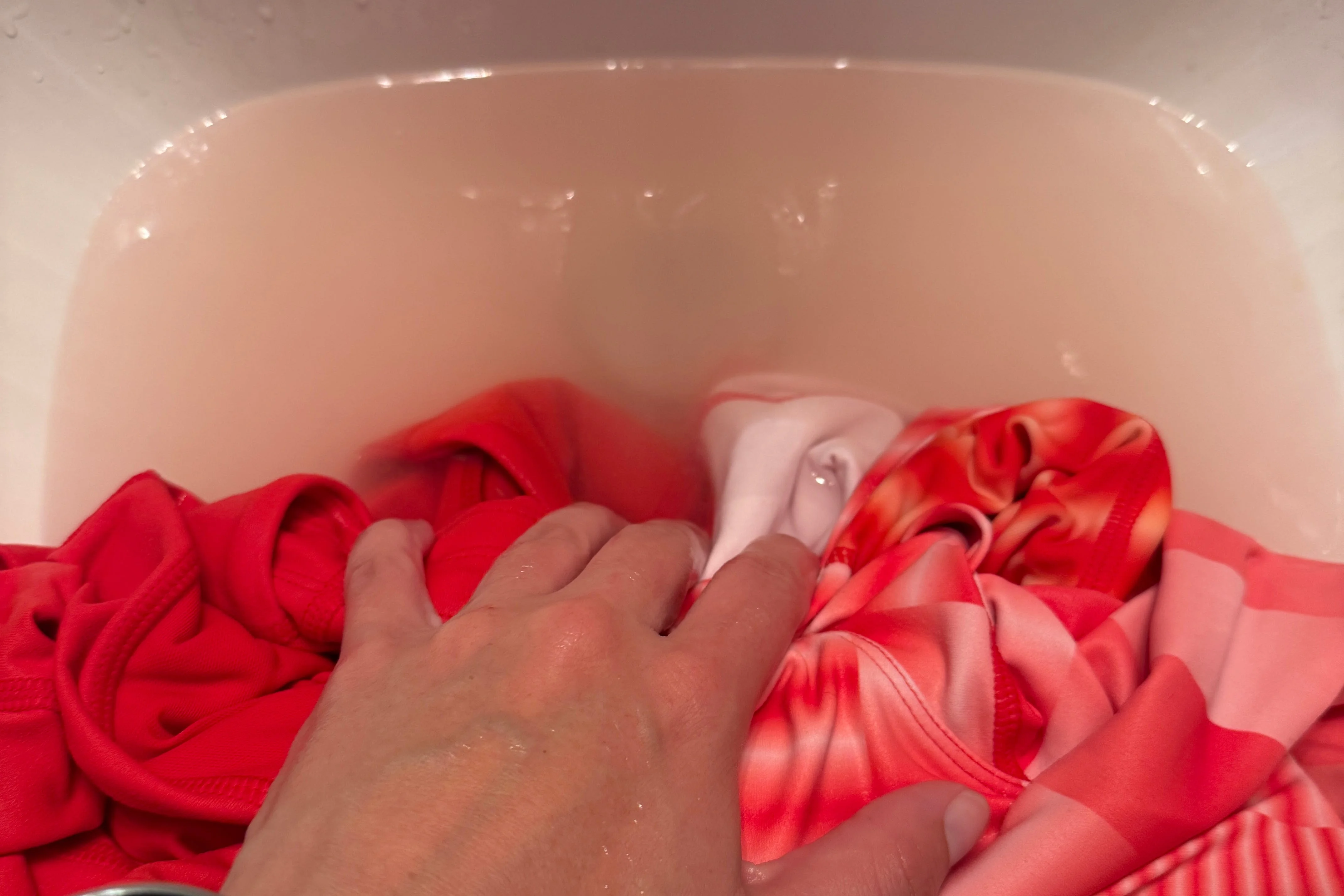 A hand submerged in a sink filled with water and red and pink clothing items. A hand submerged in a sink filled with water and red and pink clothing items.